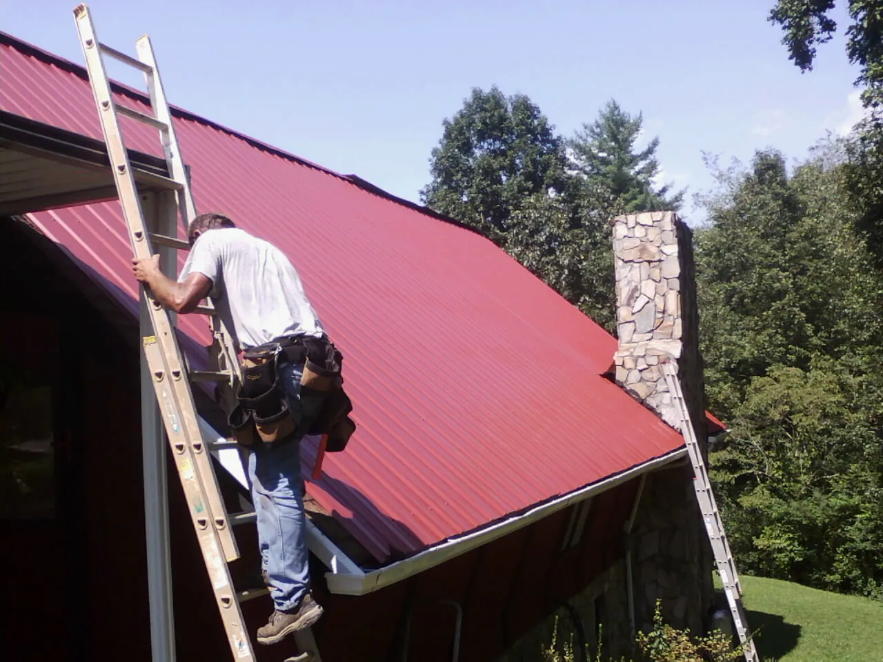 Roofer on a ladder beside a red metal roof and stone chimney — Soffit & Fascia Repair by Roofing Brothers in Masontown